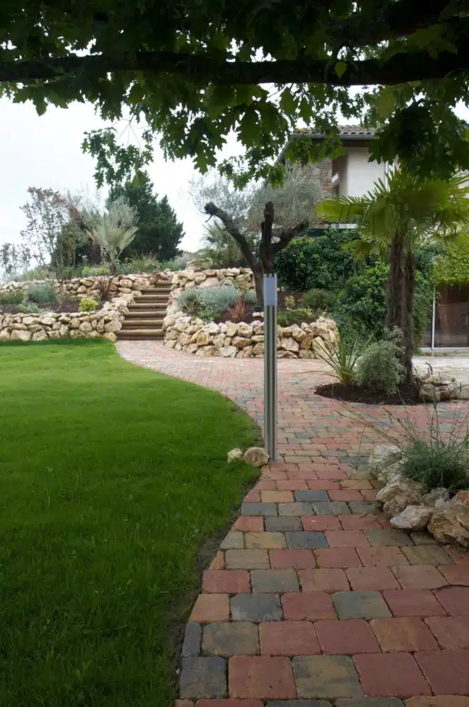 Jardin paysager avec pelouse verte, allée pavée multicolore, escalier en pierre et bois, murs de soutènement et diverses plantes.