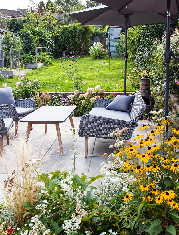 Jardin arrière luxuriant avec patio gris moderne, mobilier de salon, parasols et massifs de fleurs jaunes, blanches et roses. Espace extérieur accueillant.