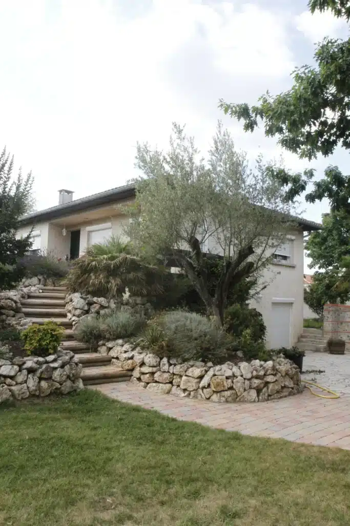 Maison avec jardin méditerranéen : olivier et terrasses en pierre Maison moderne claire avec toit gris, volets blancs. Jardin méditerranéen en terrasses, olivier, murs de pierre, escalier en bois, pelouse.
