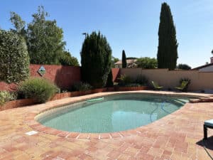 Piscine ovale carrelée de bleu avec terrasse en briques rouges, entourée de murs colorés et verdure luxuriante. Jardin ensoleillé, ciel clair.