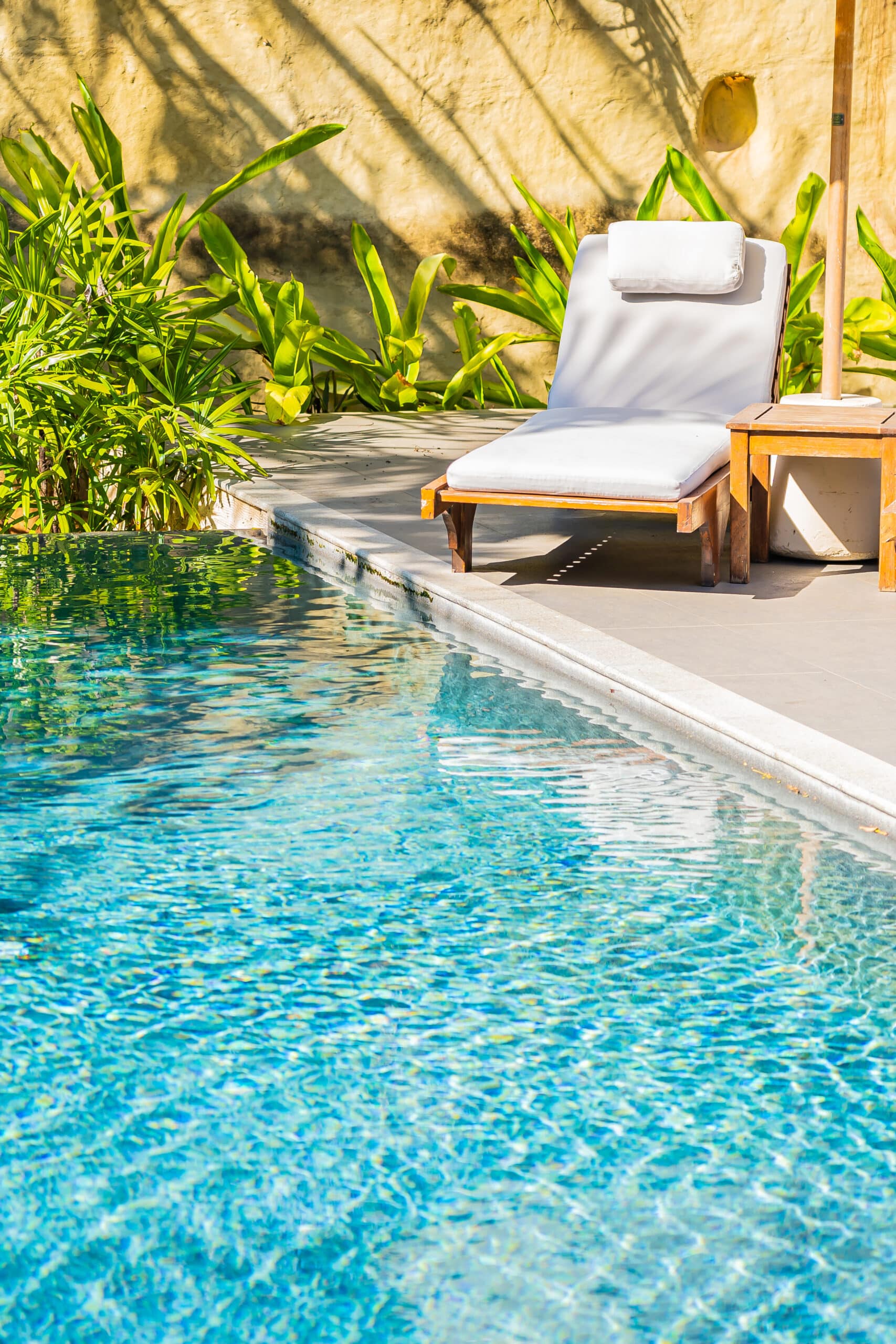Piscine bleue scintillante sous le soleil avec transat blanc et table en bois sur une terrasse, entourée de plantes tropicales.